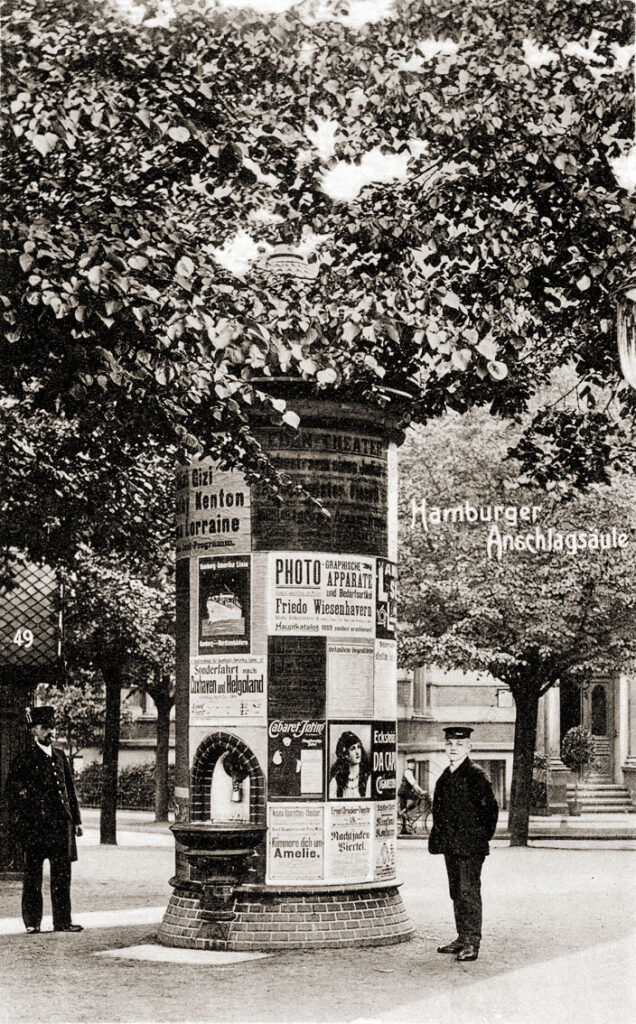 Historische Litfaßsäule mit Plakaten, zwei Männer in Anzug und Mütze, Umgebung mit Bäumen.