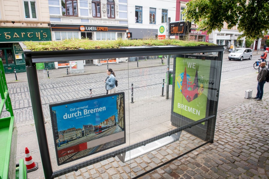 Bushaltestelle in einer Stadt mit begrüntem Dach, Plakate und Menschen im Hintergrund.