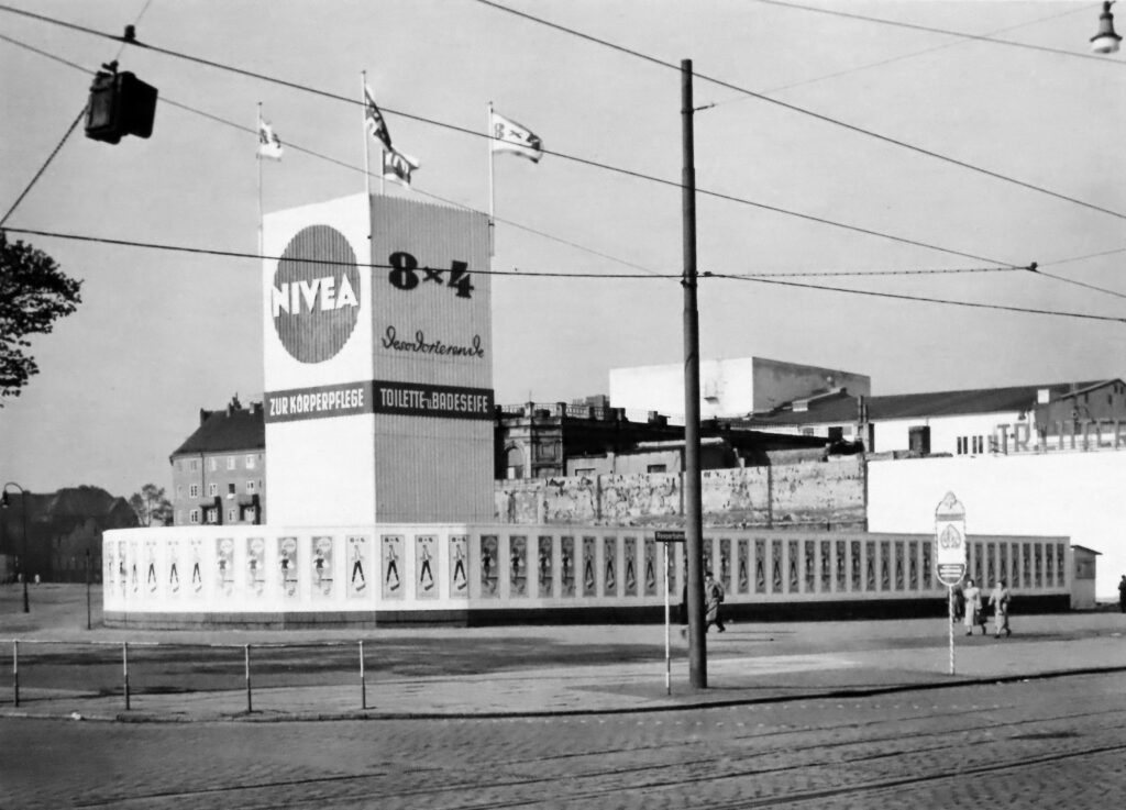 Eine Straßenwerbung für Nivea in Schwarzweiß mit Fahnen und Passanten, aus den 1930er Jahren.