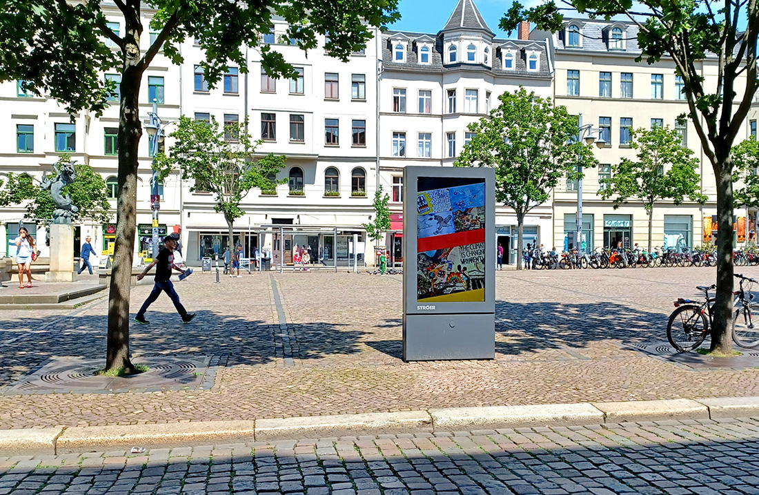 Ein belebter Platz mit Bäumen, Passant, digitaler Werbeschild, Fahrräder und historische Gebäude.