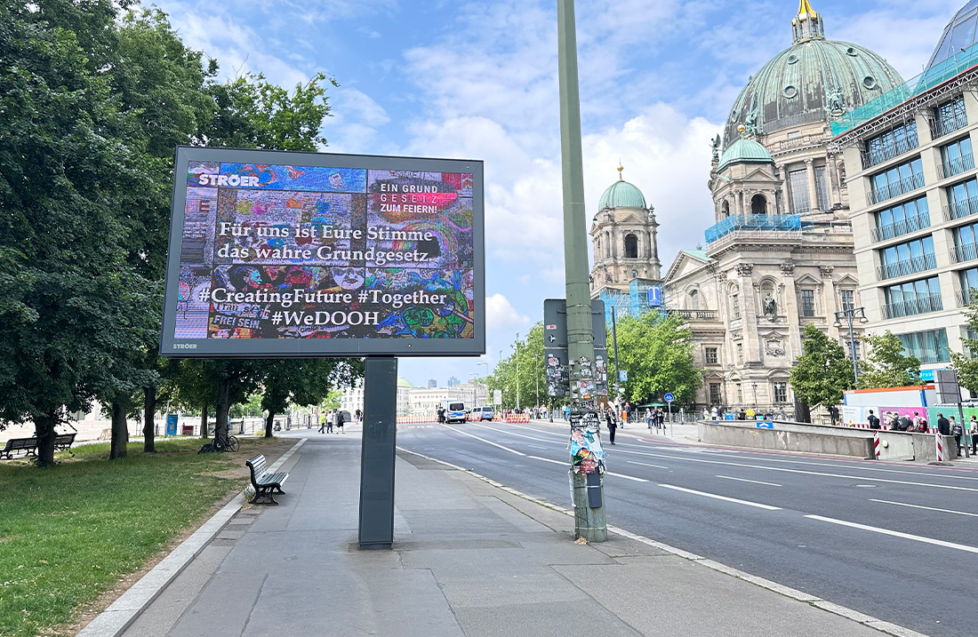 Digitale Werbetafel in Berlin, Straße, Dom, blauer Himmel, grüne Bäume, wenige Personen.