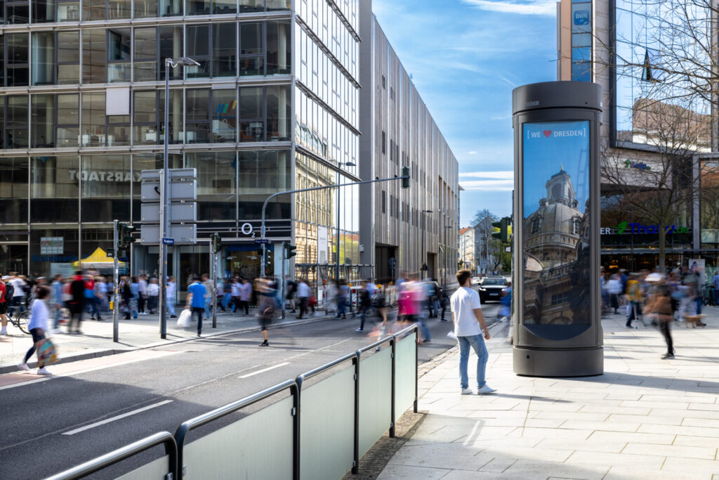 Eine belebte Stadtstraße mit Fußgängern und einem großen Werbeschild auf dem Bürgersteig.