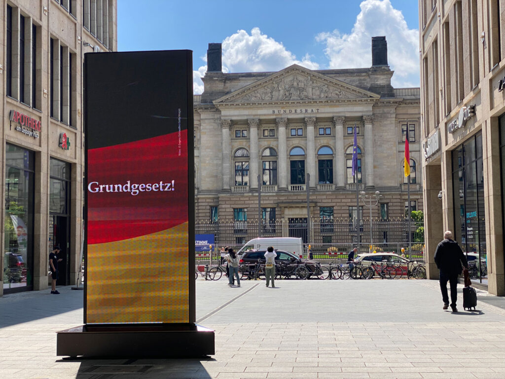 Großer Bildschirm zeigt "Grundgesetz!" in der Nähe eines historischen Gebäudes. Menschen laufen vorbei.