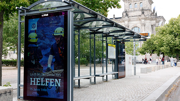 Zwei Helfer in gelber Schutzkleidung lächeln freundlich auf einem Plakat an einer Haltestelle